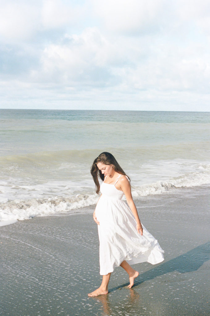 Mom walking on the beach by the waves in a white dress taken by Maternity Photographer in Charleston,SC